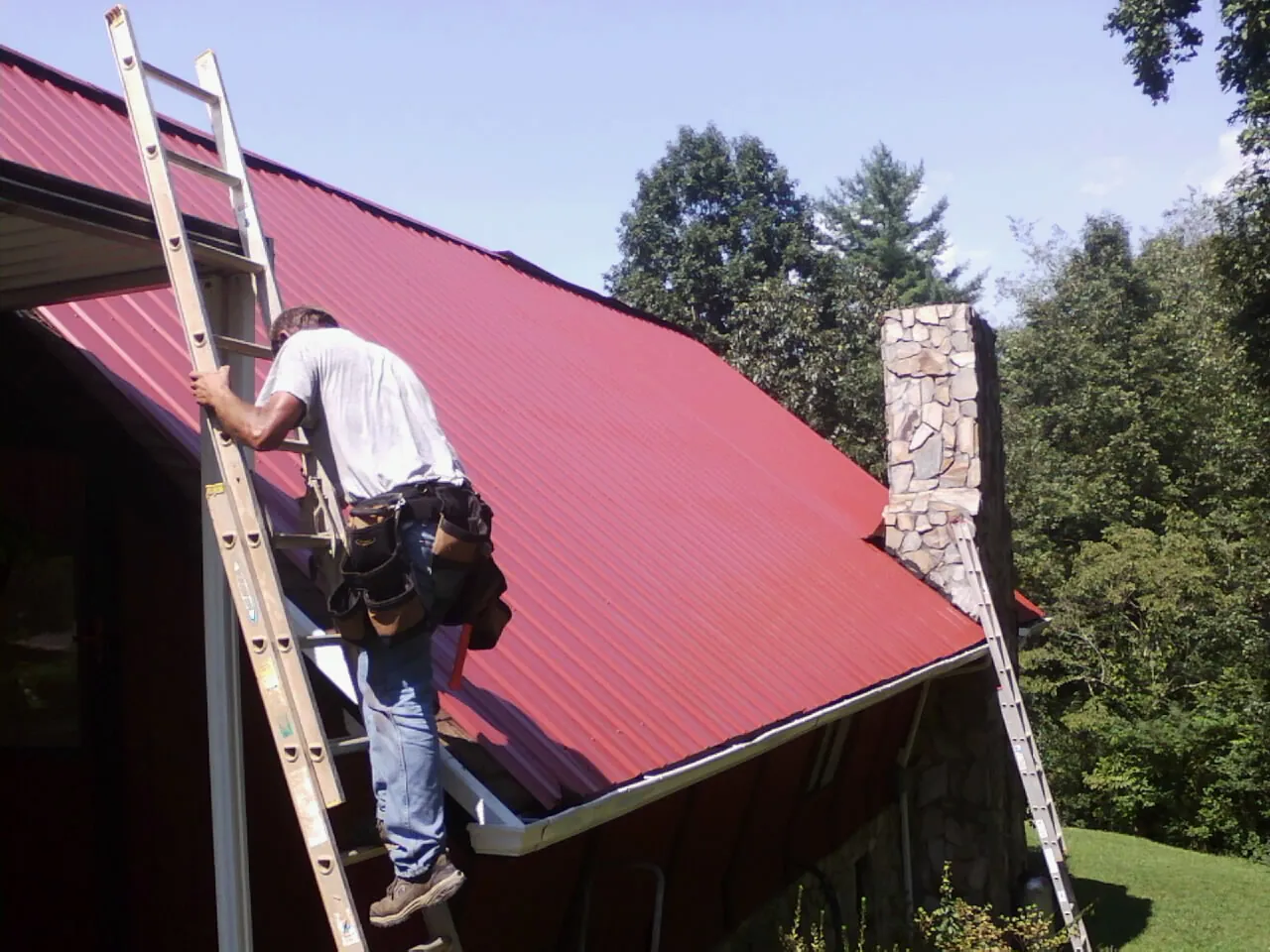 Roofer on a ladder beside a red metal roof and stone chimney — Soffit & Fascia Repair by Roofing Brothers in Westbrookville